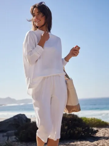 Woman wearing a white linen shirt and white linen capri pants, carrying a tote bag; standing on a beach.