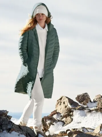 Woman wearing a green insulated parka over white clothing and hood up over a white winter hat, standing on a rocky hilltop.
