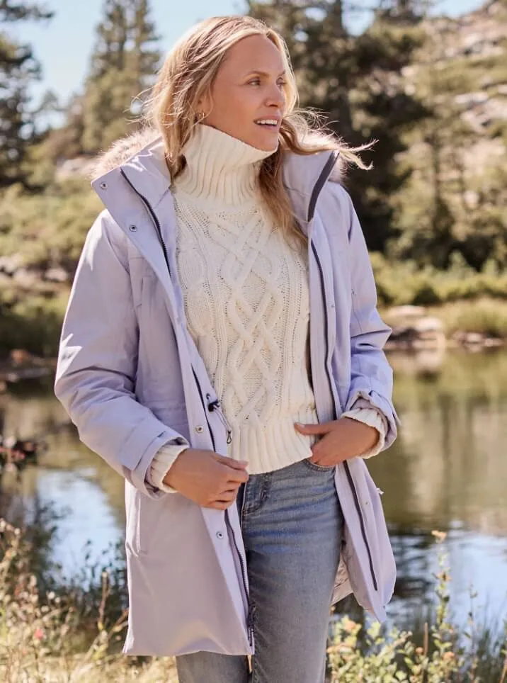 Woman wearing a lavender parka over a white cable knit turtleneck sweater and blue jeans, standing outdoors near a stream. 