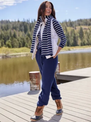 Woman wearing a navy and white long sleeve shirt with matching scarf, a white vest, navy blue sweatpants and duck boots; walking on a pier.