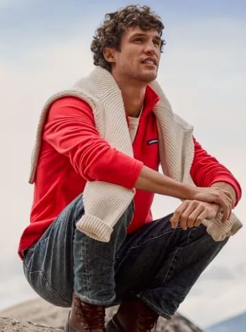 Man wearing a red quarter-zip fleece and blue jeans with a white sweater draped over his shoulders. 