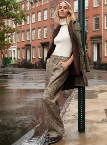 Woman wearing a white short sleeve turtleneck sweater under a maroon blazer and olive green jacket, with brown flannel wide leg pants; leaning against a street sign.
