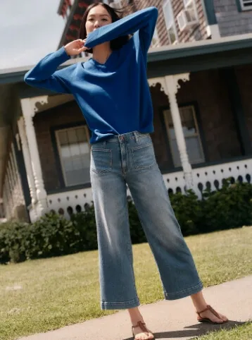 Woman wearing a blue sweater and cropped, wide legged blue jeans; standing on a sidewalk.