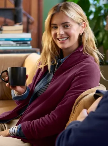 Woman wearing a maroon fleece, holding a coffee cup while seated on a couch. 