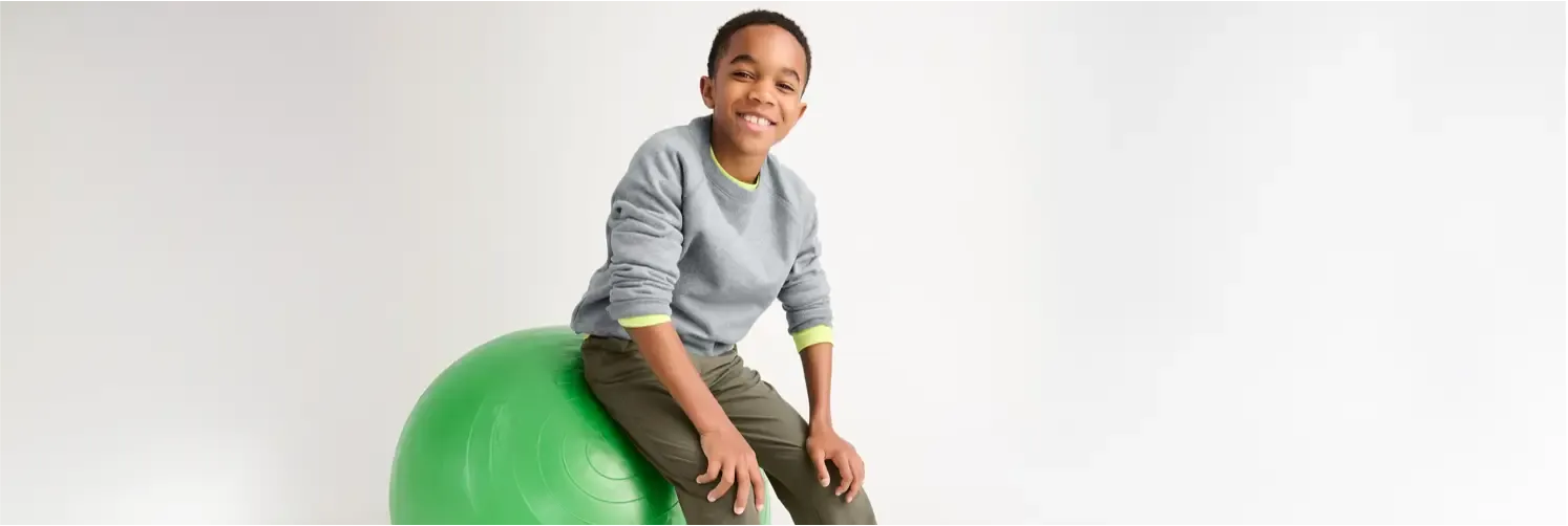 A young boy in a gray sweater with neon green cuffs sitting on a green exercise ball.