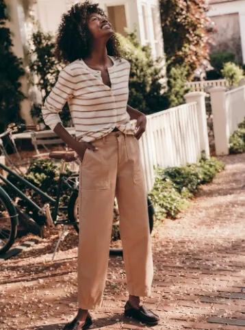 Woman wearing a white and tan long sleeve button down crew neck shirt and tan barrel leg pants; standing on the sidewalk smiling up at the sky.