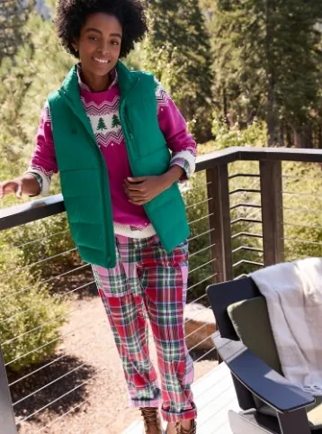 Woman wearing a pink holiday sweater under a green puffer vest, with pink and green plaid pajama pants.