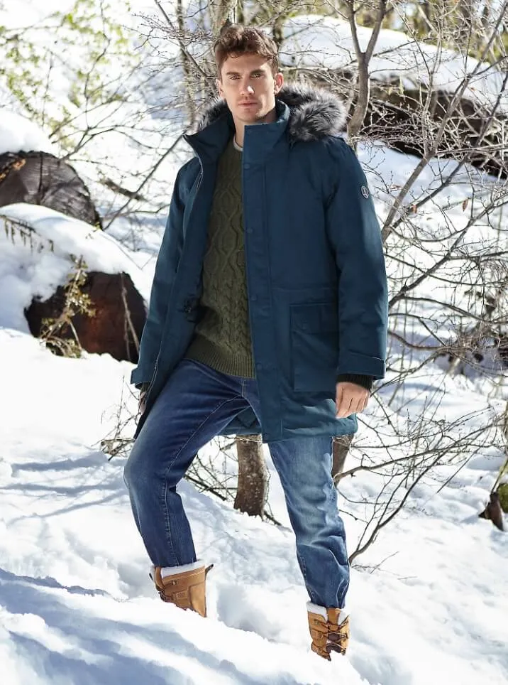 Man wearing a navy blue parka over a green cable knit sweater and blue jeans, standing on a snowy hillside.