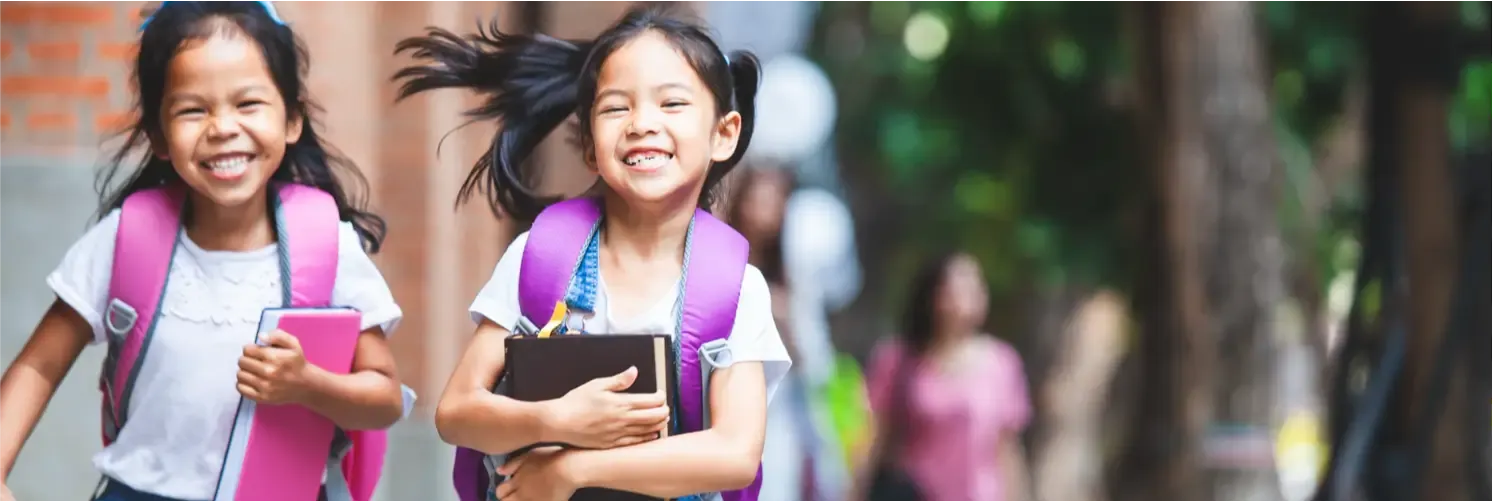 Two young girls running outdoors with pink backpacks, one holding a notebook and the other with a happy expression.
