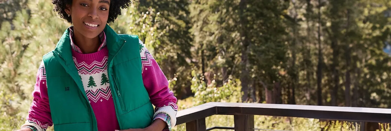 Woman wearing a pink holiday sweater under a green vest, standing outdoors.