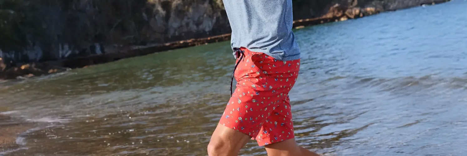 Man wearing a heather blue T-shirt with bright red floral print swim shorts walking on a beach.