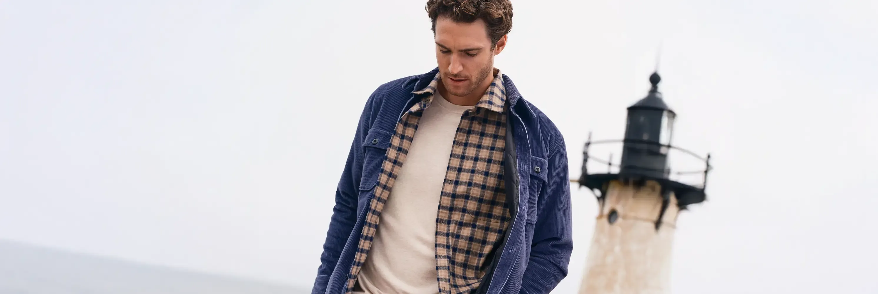 Man in a navy corduroy jacket with a plaid shirt near a lighthouse.