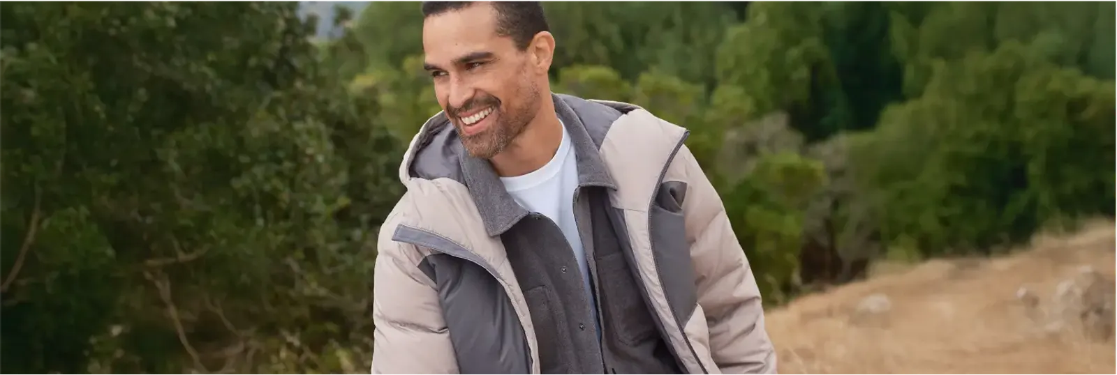Man wearing a light beige hooded puffer jacket layered over a gray zip-up and a white T-shirt.