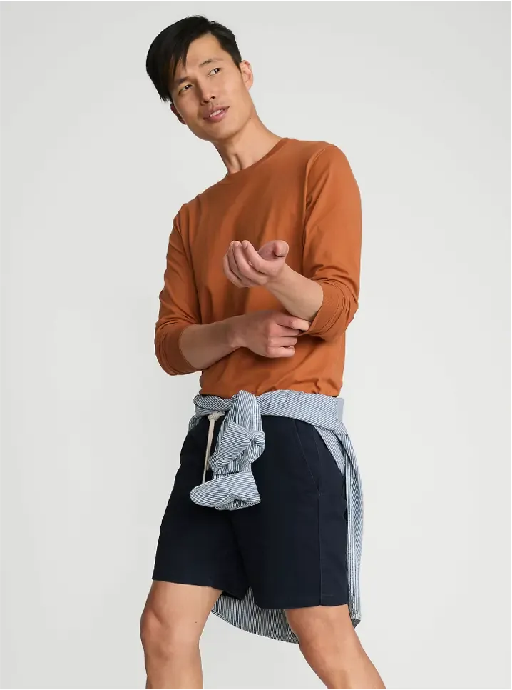 Man wearing an orange long-sleeve shirt with navy shorts, a light blue sweater tied around the waist.
