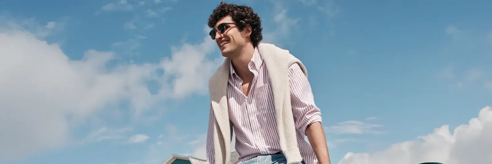 Man in sunglasses wearing a white and red striped button up shirt with a beige sweater draped over his shoulders.