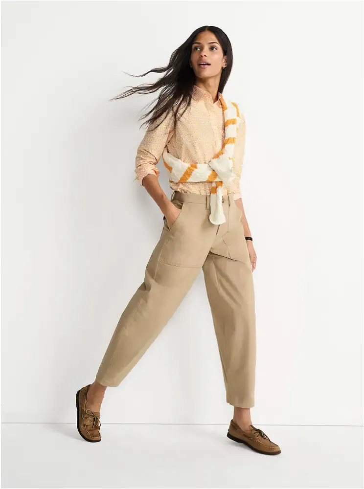 A woman in beige cropped pants and light orange button-up shirt, a white and orange striped sweater tied across her chest, and brown leather shoes.