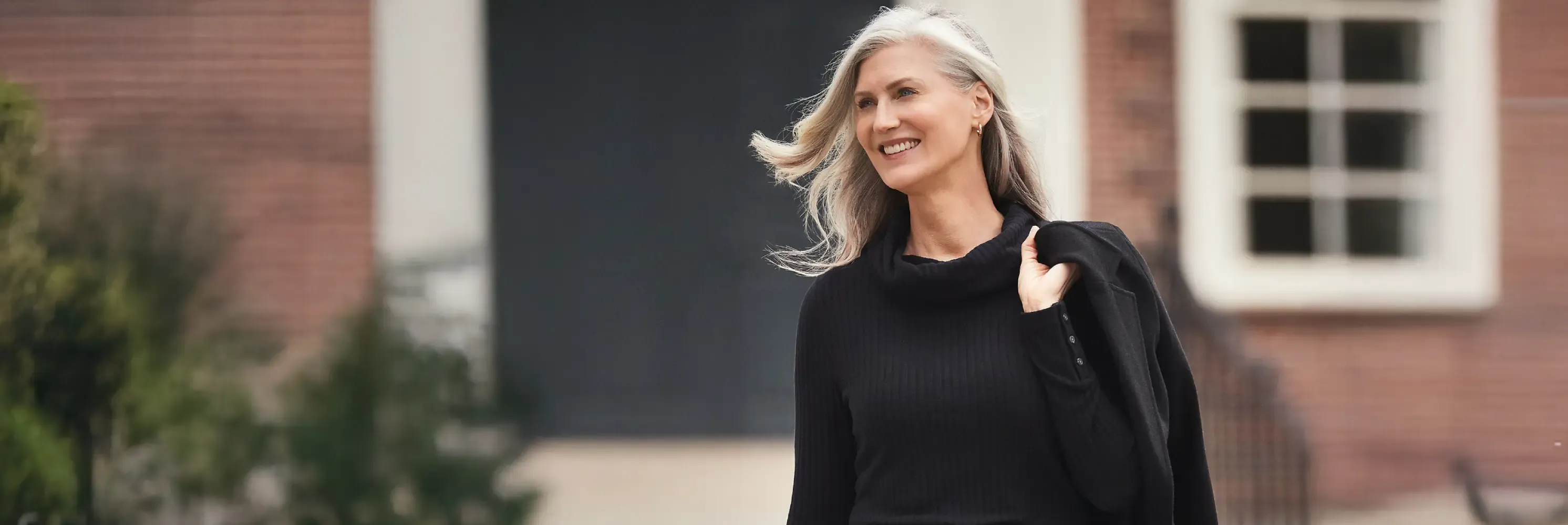 Woman wearing a black ribbed sweater dress with a blazer slung over her shoulder.