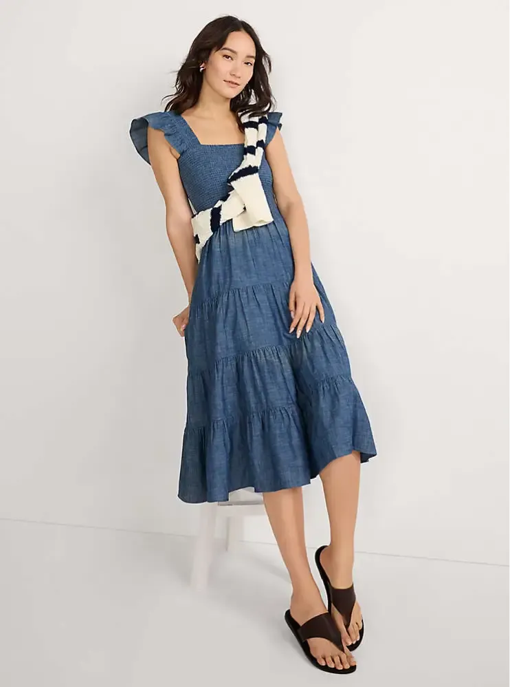 A woman wearing a blue smocked tiered dress with ruffled straps, a striped sweater draped over her shoulder, and wearing black sandals.