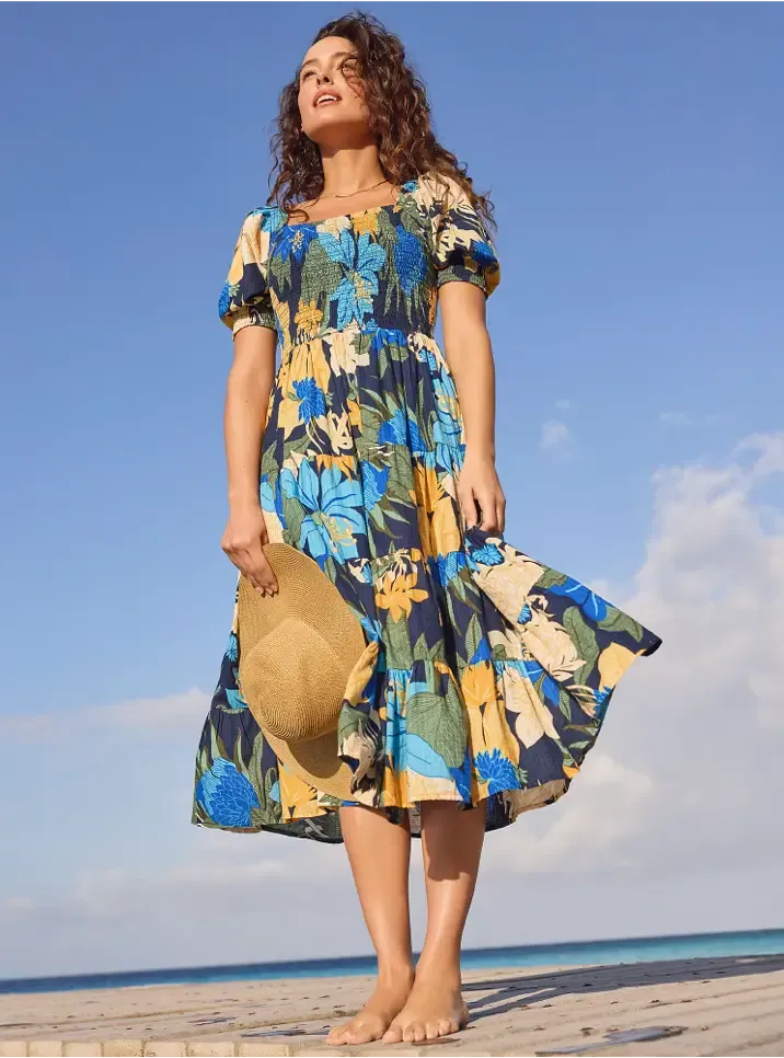 Woman with curly hair wearing a vibrant blue and yellow floral midi dress, barefoot, holding a straw hat, standing on a wooden boardwalk near a beach.