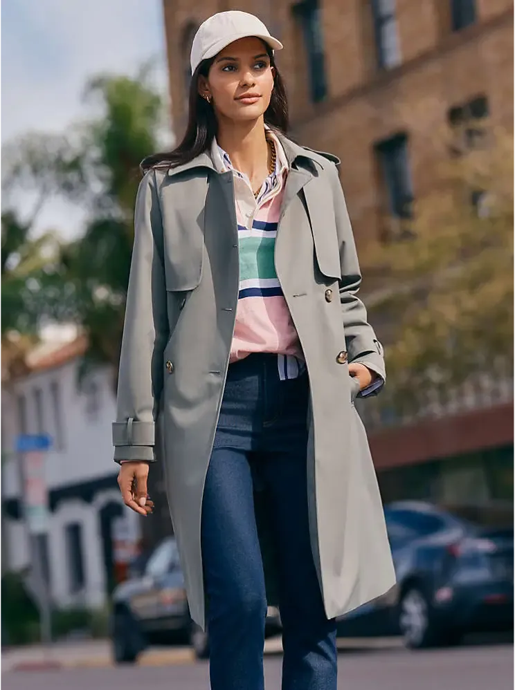 Woman wearing a light gray trench coat over a pink and blue striped shirt and dark blue jeans.