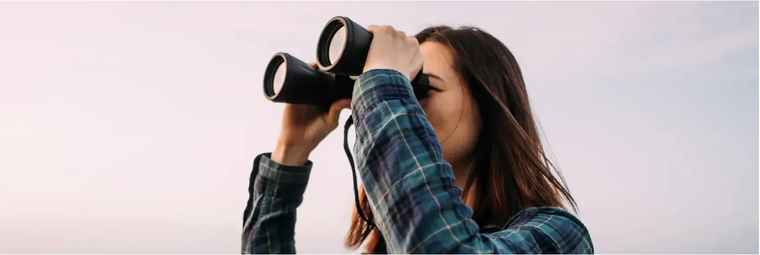Woman wearing a blue and green plaid shirt, looking through binoculars against a clear sky background.