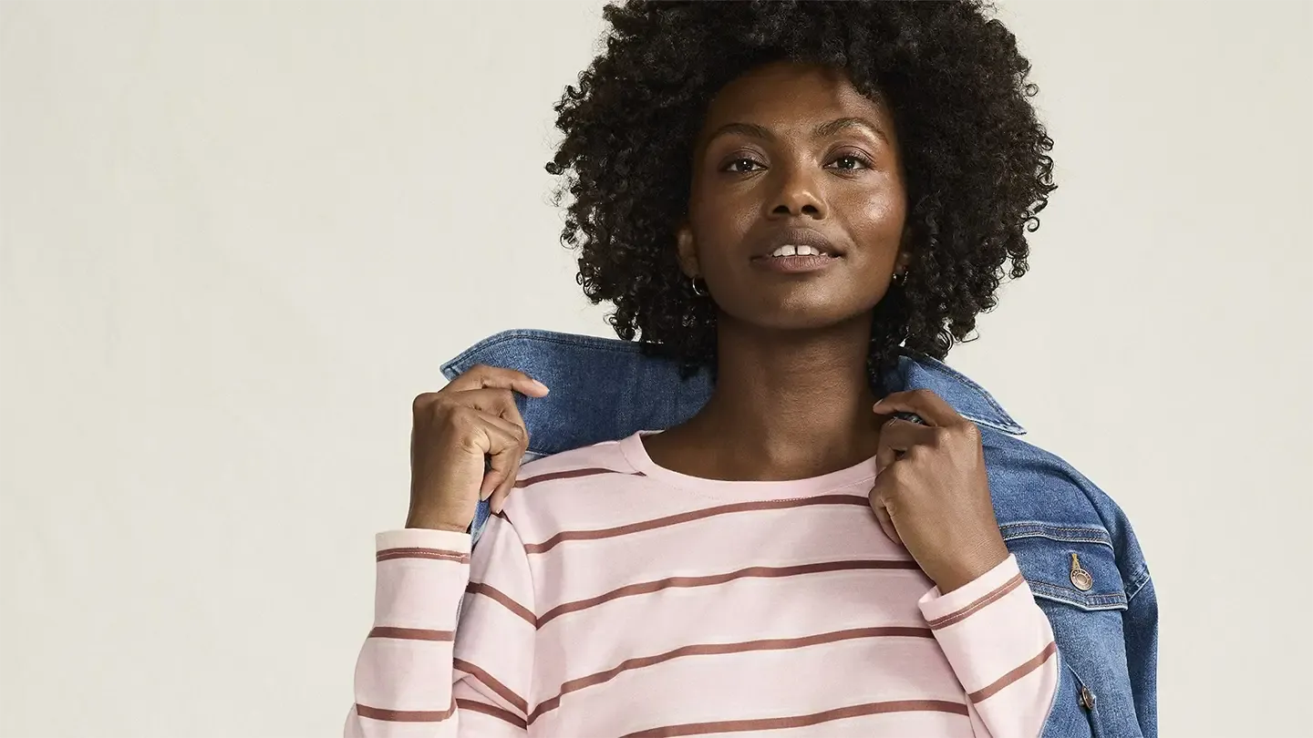 Woman wearing a pink and brown striped long sleeve crew neck pulls a jean jacket over her shoulders.