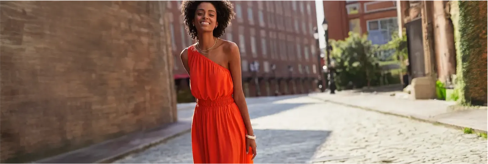 Woman wearing a bright red-orange one-shoulder midi dress walking down an urban street.