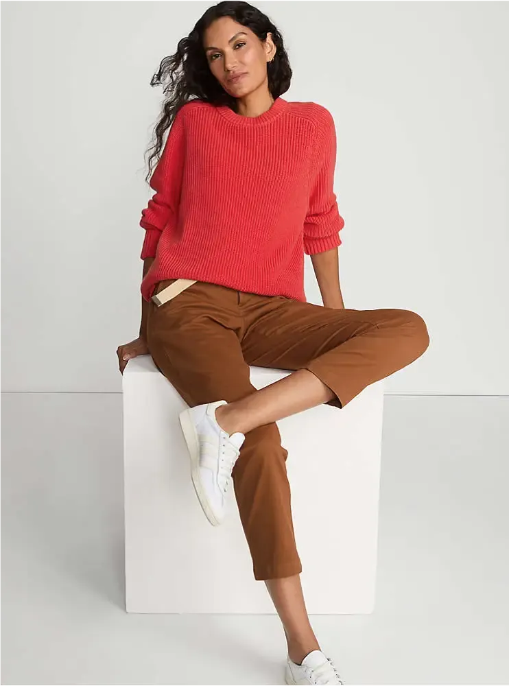 Woman sitting on a white block and wearing a bright red knit sweater and brown cropped chinos.