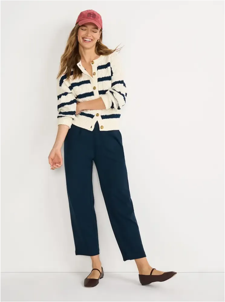 A woman with brown hair wearing a white and navy striped cardigan, navy pants, brown flats, and a pink baseball cap.