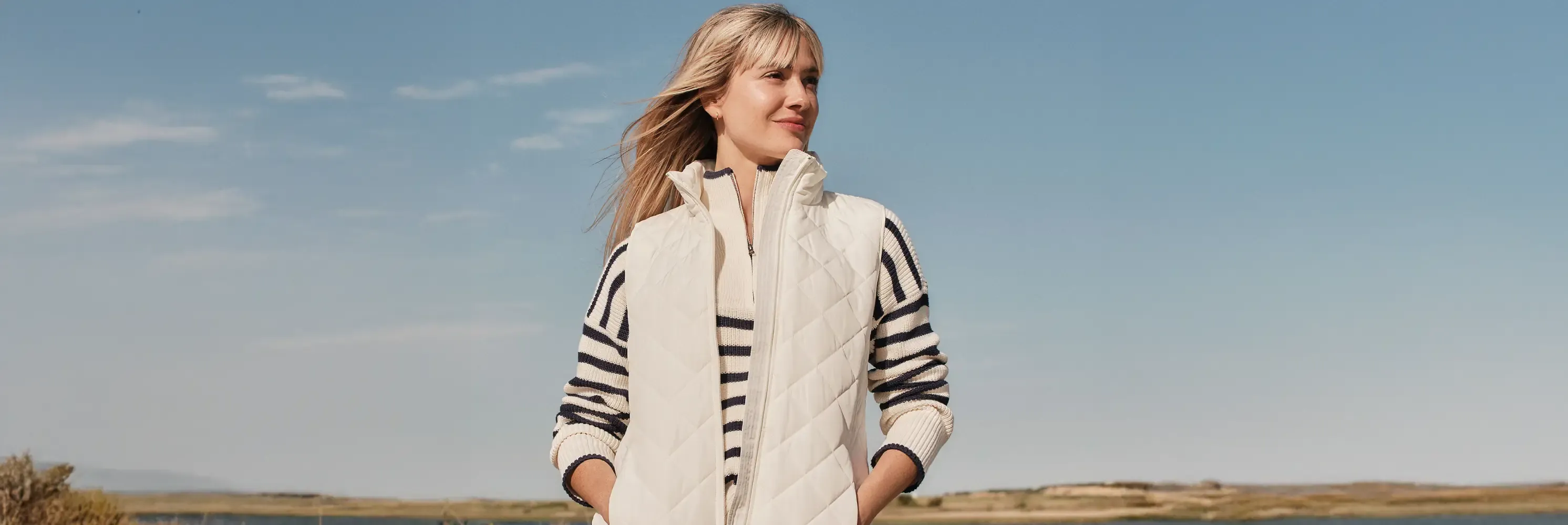 Woman in a cream quilted vest over a striped sweater standing in an open field.