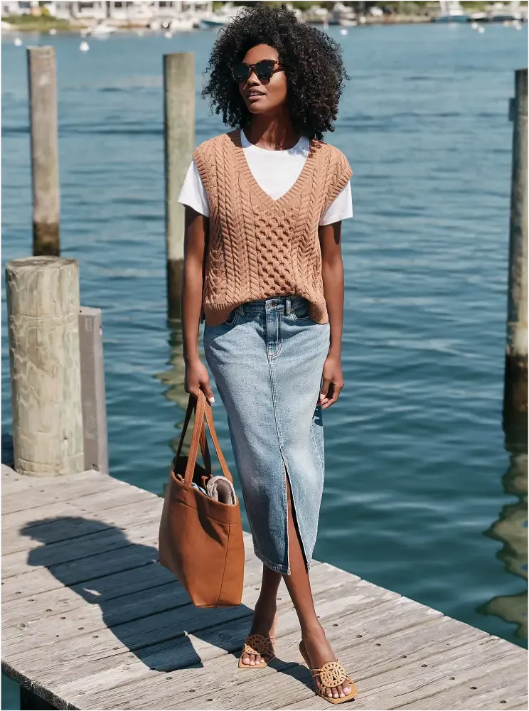 Woman wearing a tan cable-knit vest over a white T-shirt, and a denim midi skirt.